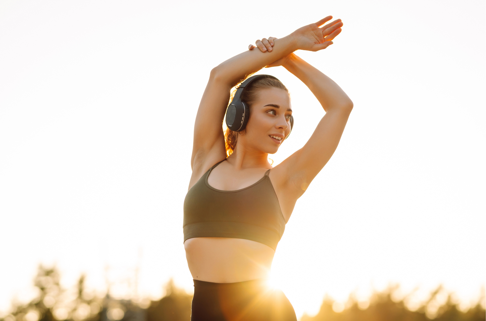Девушка в наушниках выполняет растяжку рук на фоне заката